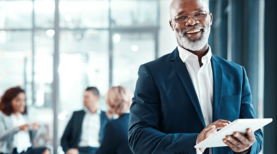 Portrait of a mature businessman using a digital tablet in a modern office with his colleagues in the background.