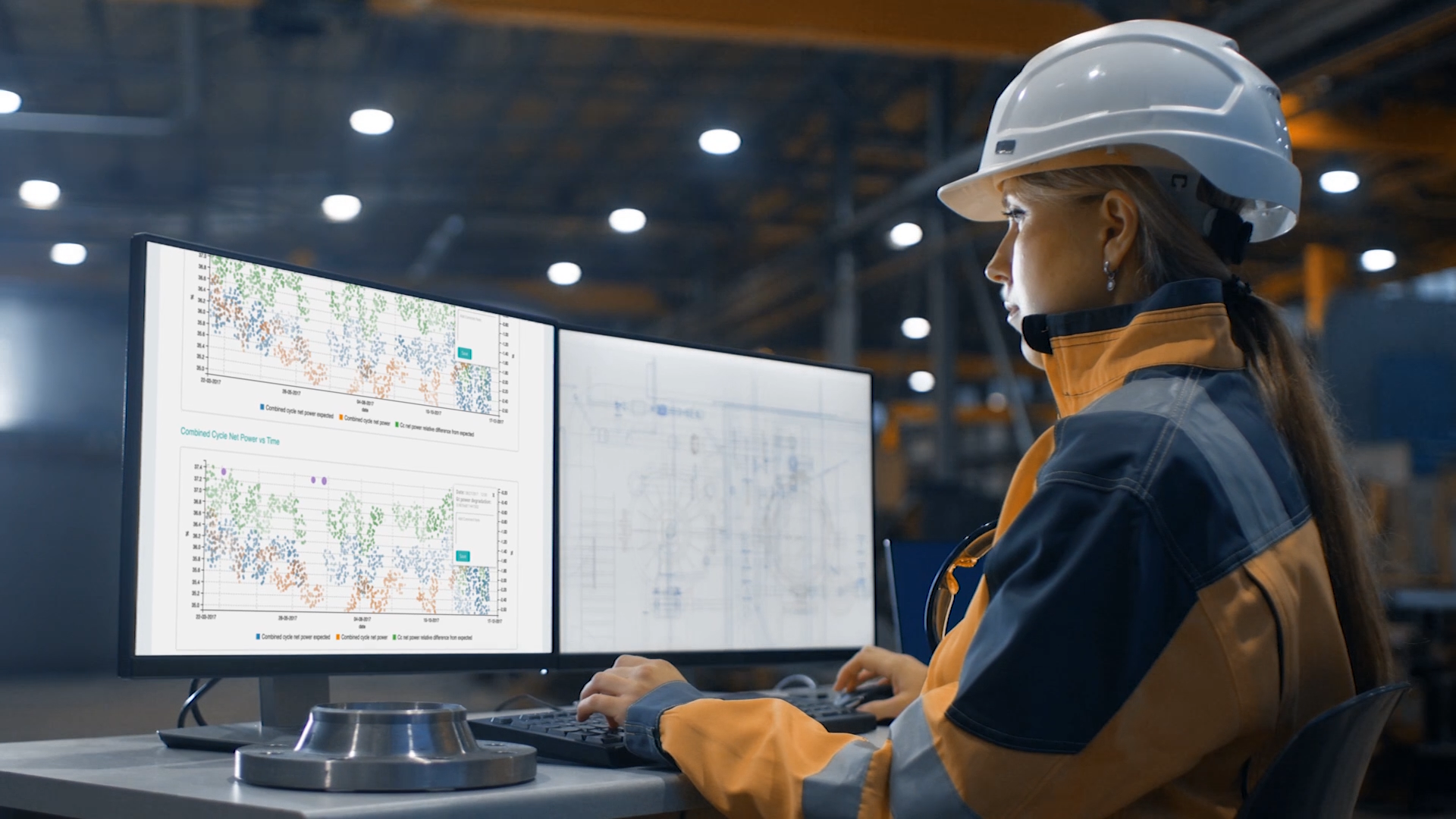 Woman with safety vest and helmet working at a desk with dual monitor system