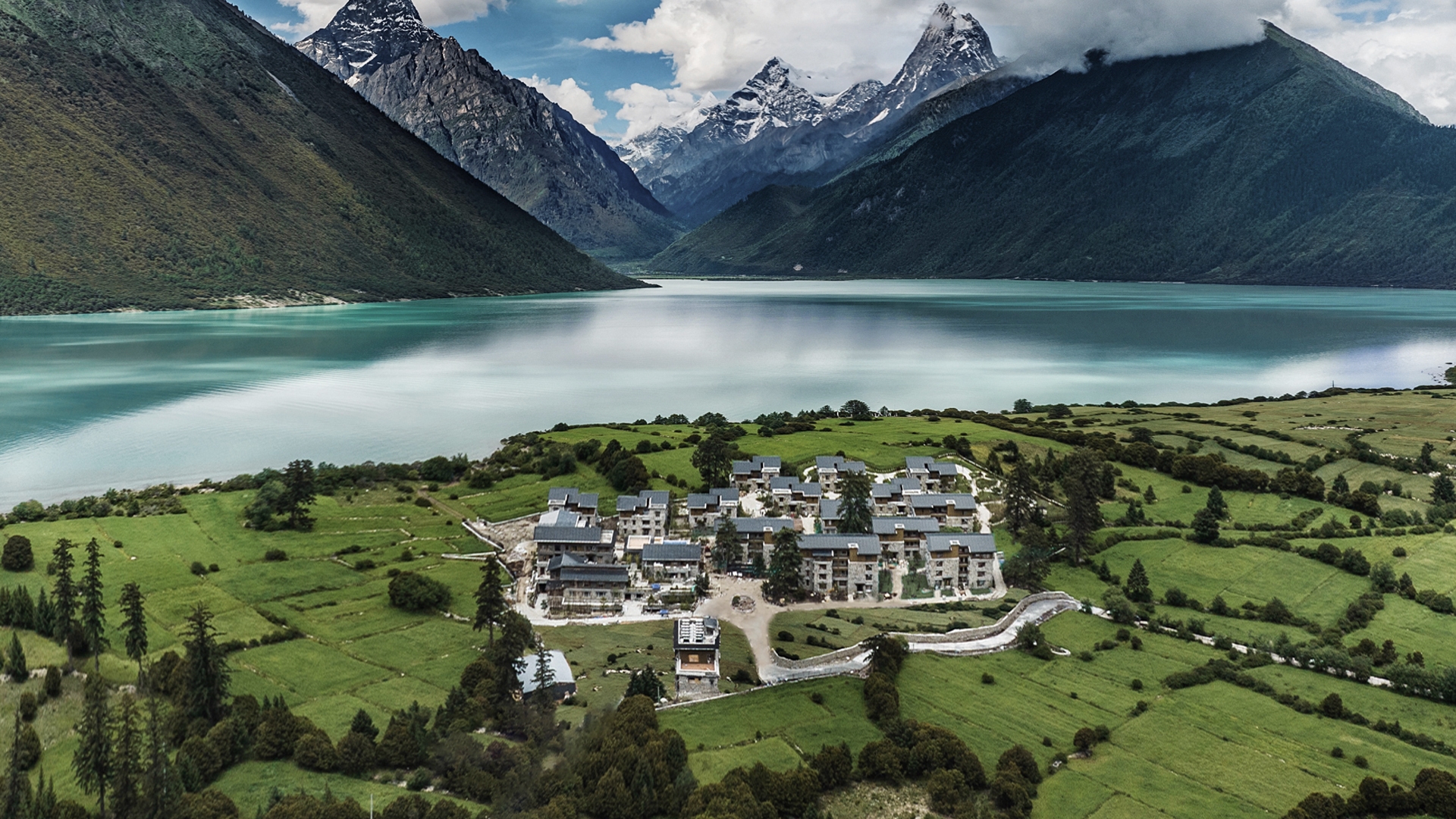 Aerial view a hotel surrounded by green fields with a lake and mountains in the background