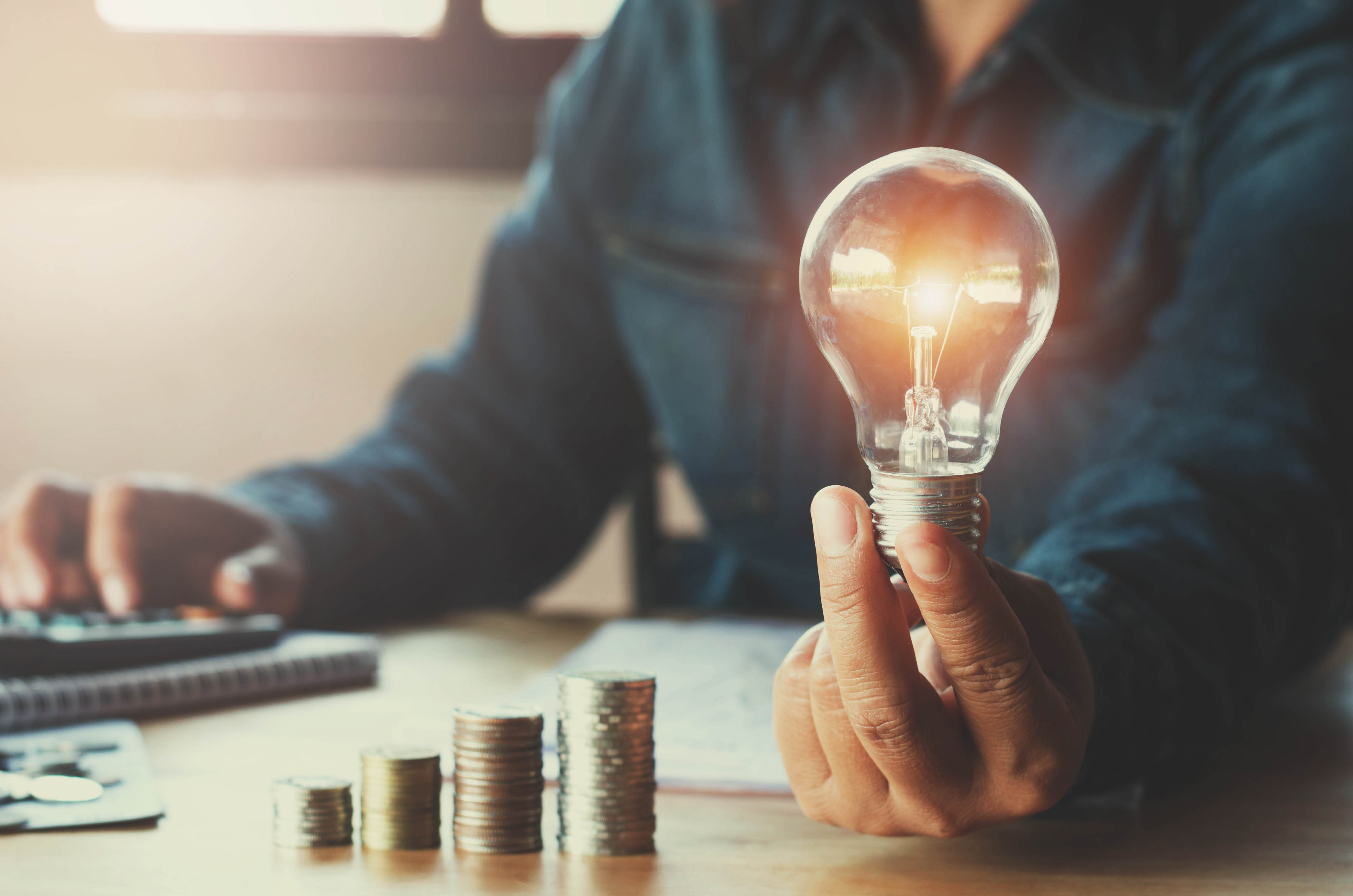 Man holding a lightbulb with a stack of coins next to him symbolizes the economic benefits offered by hybrid power solutions in remote locations