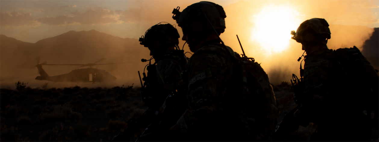 Silhouetted, sunrise view of three soldiers in tactical gear and helmets walking past a grounded helicopter in the background